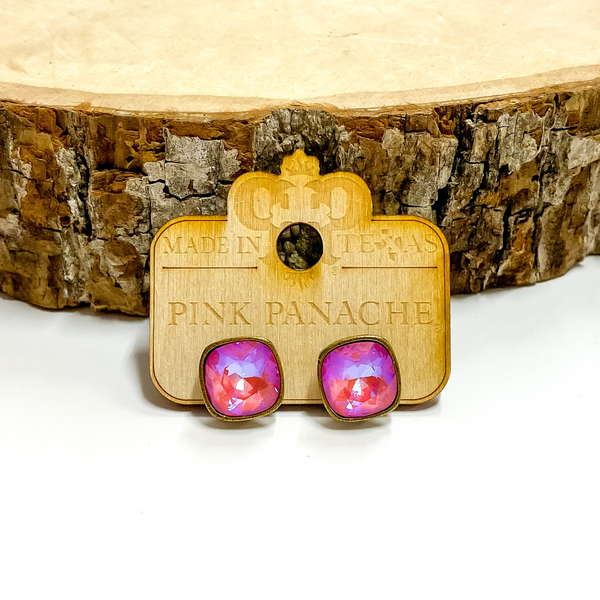 A pair of bronze, square stud earrings with pink lotus delight cushion cut crystals. These earring are pictured on a wood holder on a white background with a piece of wood in the background.