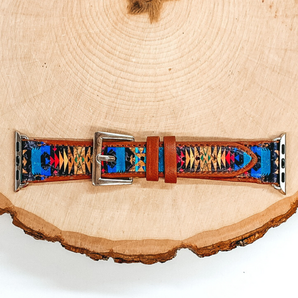 Tan watch band with an Aztec design inlay in multicolored. This watch band has a silver clasp and silver Apple watch band accessories. This watch band is pictured on a piece of wood on a white background.
