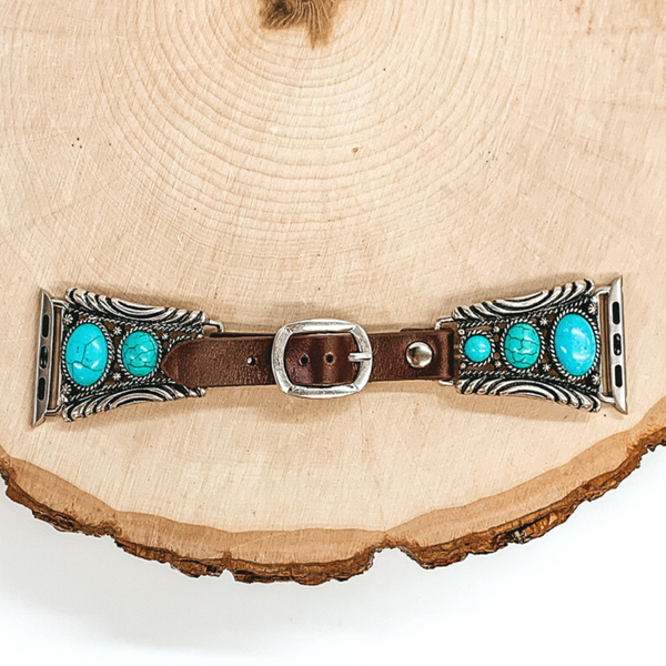 Dark Brown watch band with silver, trapesoid shaped ends with Apple watch band acessories. The ends have three turquoise stones each going from biggest to smallest. This watch band is pictured on a piece of wood on a white background.