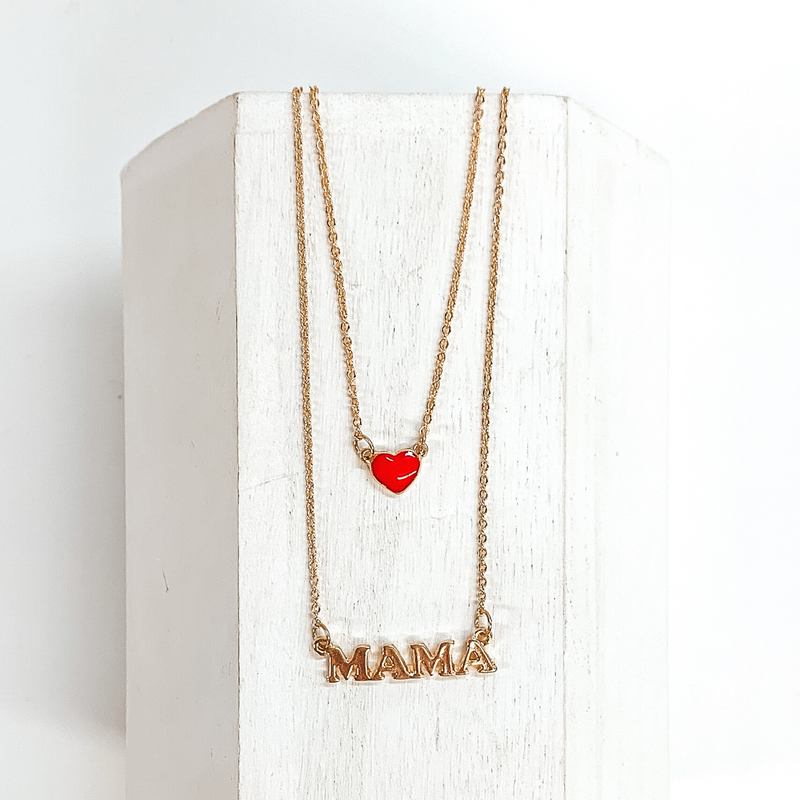 Two stranded gold chained necklace. The longer strand has a gold pendant that spells out "MAMA" and the shorter strand has a small red heart charm. This necklace is pictured laying on a white block on a white background. 