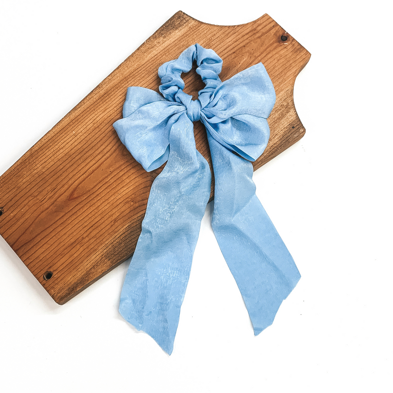 This is a light blue satin scrunchie with a long bow, this scrunchie is taken on  a brown necklace board and on a white background.