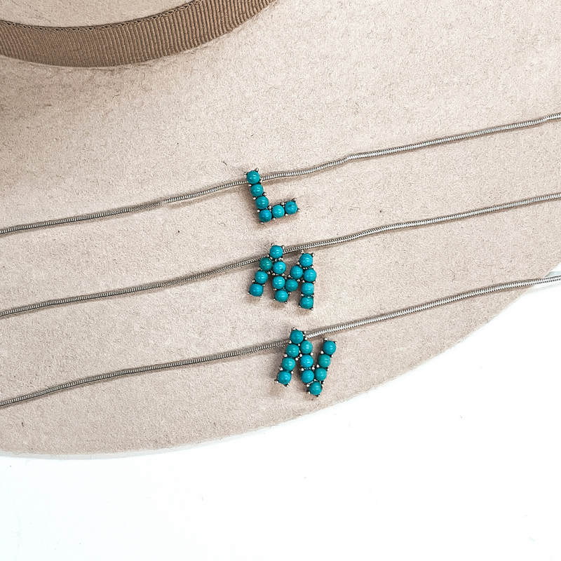 These are three turquoise stone initial necklaces with a thin, silver  snake chain. From top to bottom; L, M, N. These necklaces are taken  laying on a beige felt hat and on a white background.