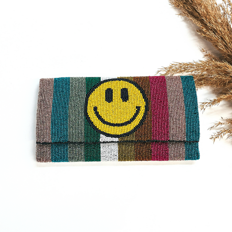 This is a seedbeaded clutch purse in multicolor with  lines and a yellow happy face. Multicolor lines in teal, rose gold, fuchsia, copper,  yellow, white, green, and mint. This bag  is taken laying on a white background with a brown plant in the side as decor.