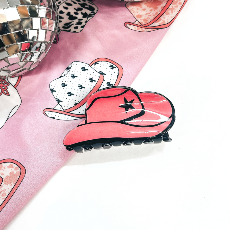 This is a pink colored cowboy hat hair clip with a black star, This clip is laying on a white background and a hat print fabric with disco balls in the back as decor.