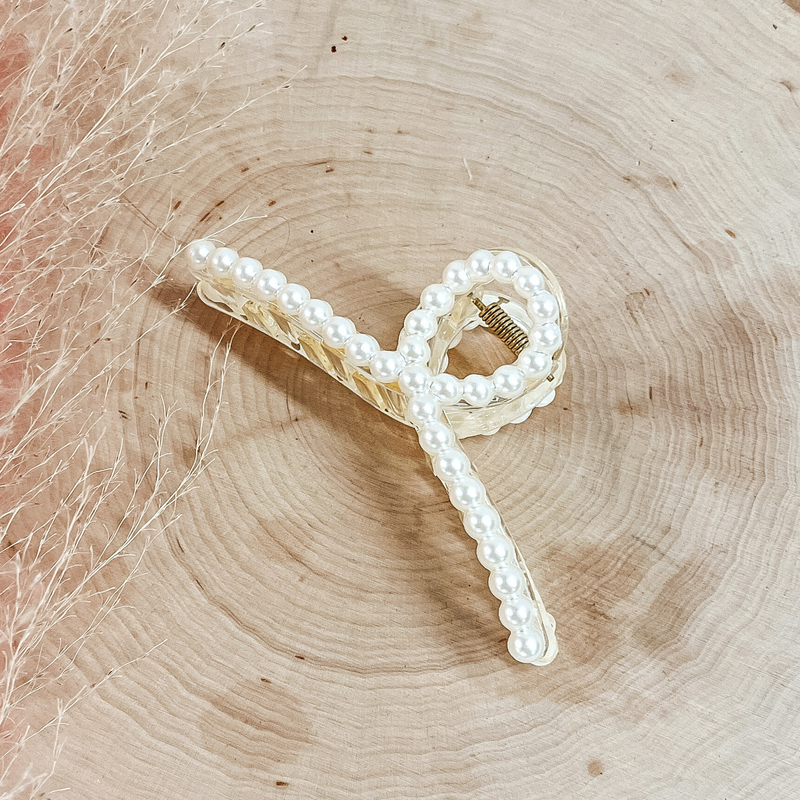 This is a long clear loop hair clip with pearls all around. This clip is laying on a slab of wood with a pink plant in the side as decor.