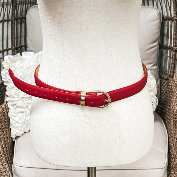 There is one skinny belt in dark red/burgundy with a small oval buckle in gold. The belt  is placed on an ivory mannequin.