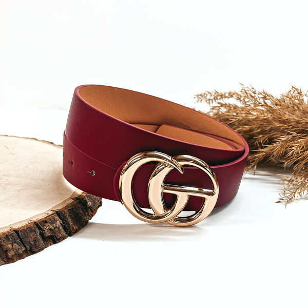 This is a burgundy/dark red-purple rolled up belt with a gold buckle in the center. This belt  is placed on a slab of wood and on a white background, there is a brown plant  in the back as decor.