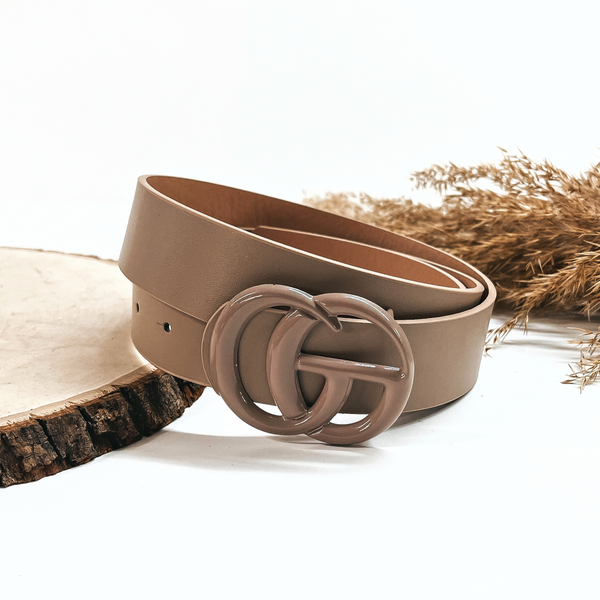 This is a taupe/brown rolled up belt with a taupe/brown buckle in the center. This belt  is placed on a slab of wood and on a white background, there is a brown plant  in the back as decor.