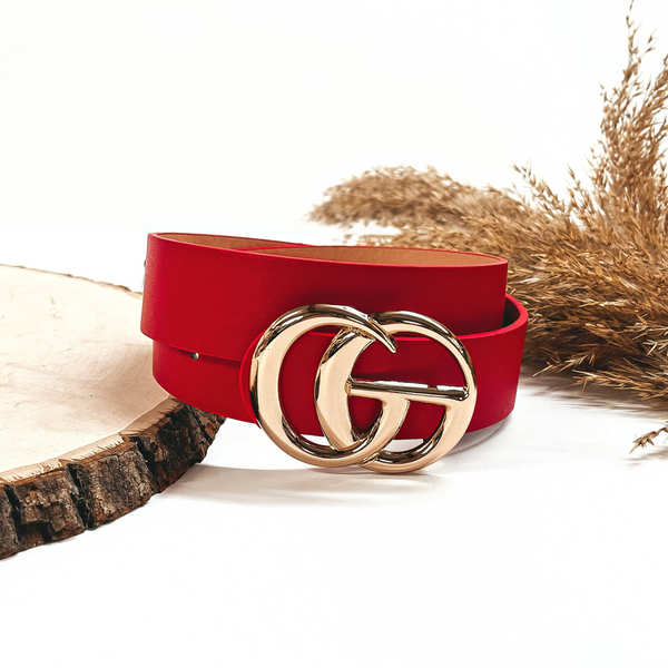 This is a red rolled up belt with a gold buckle in the center. This belt  is placed on a slab of wood and on a white background, there is a brown plant  in the back as decor.