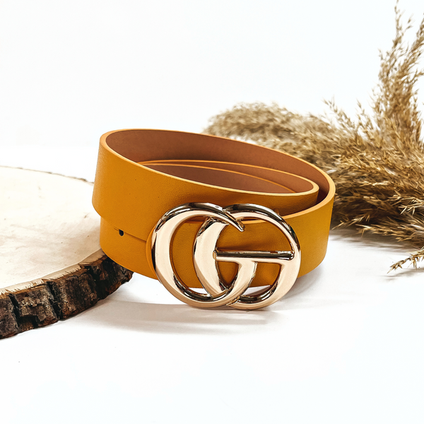 This is a yellow mustard rolled up belt with a gold buckle in the center. This belt  is placed on a slab of wood and on a white background, there is a brown plant  in the back as decor.