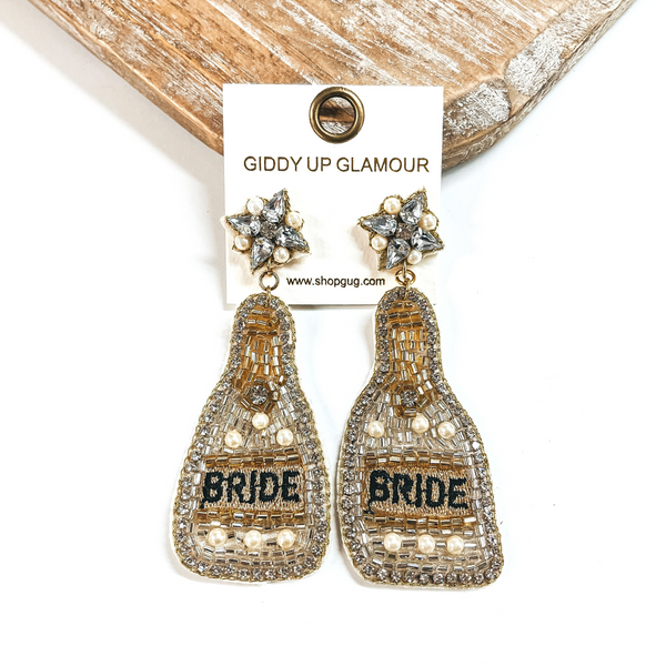 These are beaded bottle earrings in silver mix with pearls. The postback  has clear crystals and pearls with gold stitching around. The bottle drop has  clear rhinestones all around with silver, gold, and pearl beads. In the  center it says, Bride, in black and in a gold background. These earrings are  taken on a white background and leaning against a light wood slab.