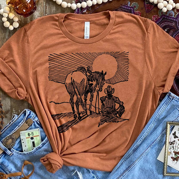 A light brown short sleeved shirt with a centered black and white graphic of a cowboy and a horse watching the sunset. Item is pictured on a brown background.