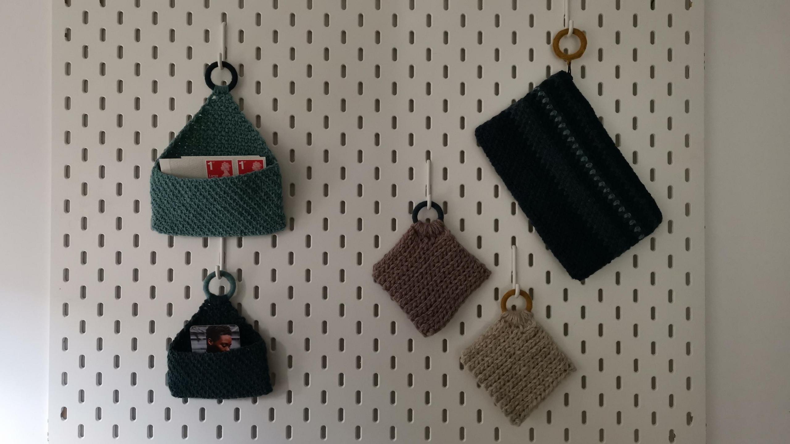 Crocheted hanging storager for stamps and business cards, a clutch and coasters are hanging from an IKEA office wall storage panel.