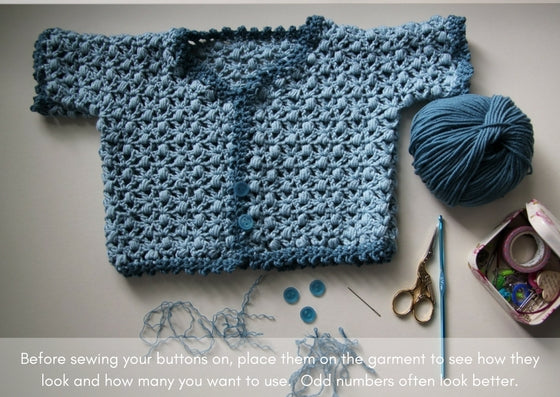 Baby cardign laid out beside a notions tin, scissors, needle and crochet hook.  Buttons are waiting to be sewn on to finish the item off.