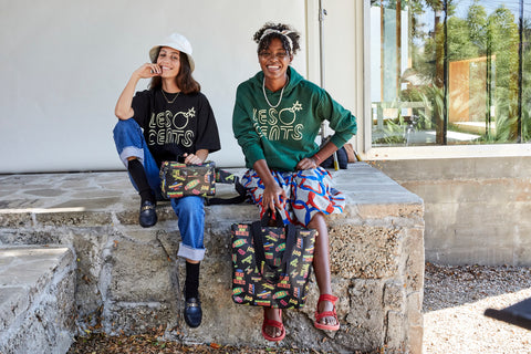 Two Models Sitting Next to Each Other and Wearing Pieces From Our Collaboration - Tee, Hooded Sweatshirt, Fanny Pack, and Tote