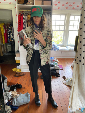Clare Vivier Taking A Selfie In Front of A Mirror, Wearing Black Pants, A Printed Green Jacket, Printed T-Shirt Underneath, and Our Ciao Green Trucker Hat