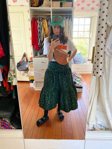 Clare Vivier Taking A Selfie In Front of A Mirror, Wearing 
 A Dark Green Jaguar Satin Skirt with Ruffles with a Printed T-Shirt and Our Grande Fanny in Natural Woven