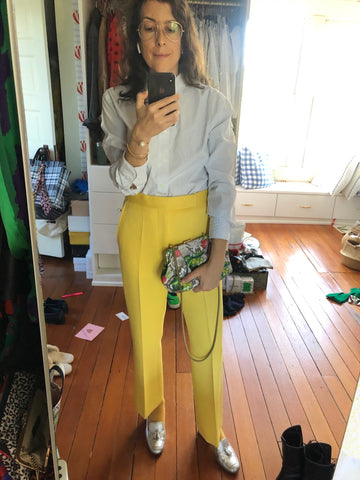 Clare Vivier Taking A Selfie In Front of A Mirror, Wearing A Light Blue Blouse with Yellow Pants and Holding Our Strawberry Snake Sissy