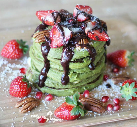 A stack of vegan matcha tea pancakes topped with strawberries and desiccated coconuts and drizzled with chocolate sauce