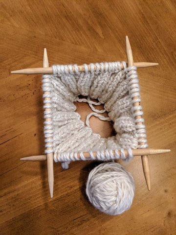 Photo of the beginning of a knit hat on four double pointed needles in a white bulky yarn.