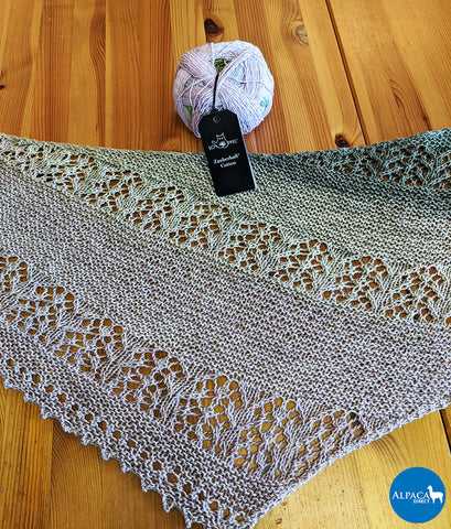 Photo of a finished Nurmilintu lace shawl with a ball of Schoppel-Wolle Zauberball Organic Cotton above it. 