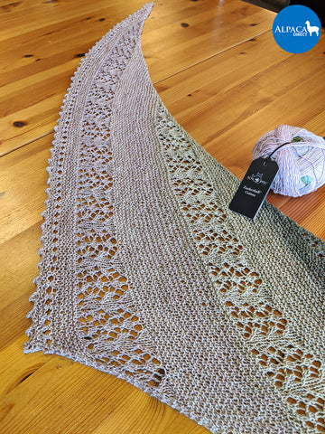 Photo of a finished knit Nurmilintu lace shawl stretched out on a wood table next to a ball of Schoppel-Wolle Zauberball Organic Cotton yarn.