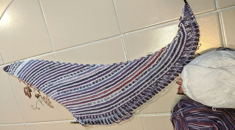 Horizontal photo of a partially knit Baubles Shawl by Andrea Mowry in the center. Two balls of Berroco Comfort Sock are to the bottom right in a solid white and purple multi color. 
