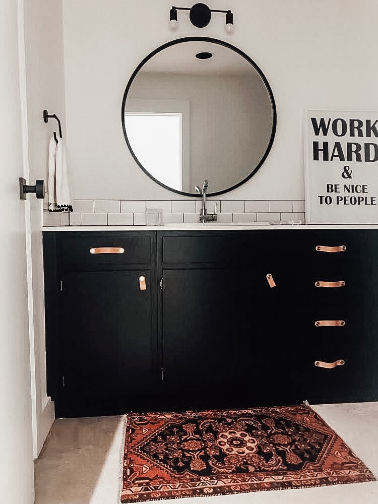 Cristina Bathroom design with a vintage rug from Swoon Rugs black cabinets, leather pulls, a round mirror, white walls