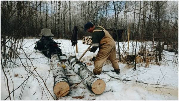 A man in Carhart Overalls, sawing felled aspen beside his Toboggan Sled