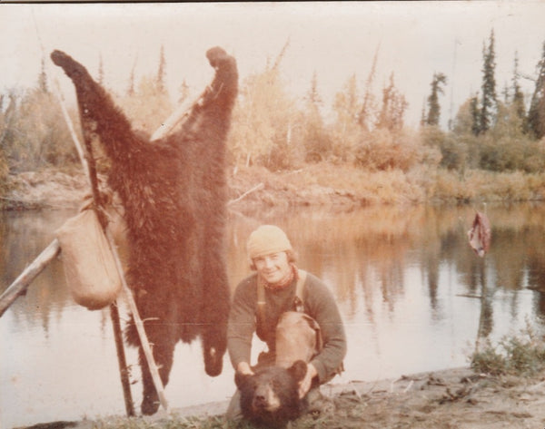 faded photo of young man in tan overalls crouching on a lake. Bearskin strung up behind him