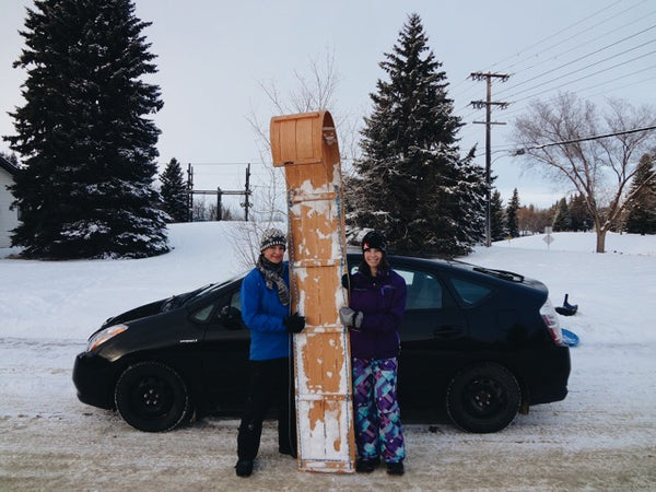 Two women in snowgear hold their upright toboggan sled beside a Toyota prius