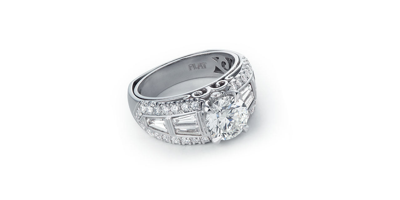 A platinum and diamond wide wedding ring with a round center diamond and a baguette diamond band bordered by round diamonds. A script initial M is in the undergallery.