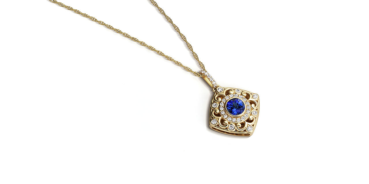 A photograph of a yellow gold Tanzanite pendant with a soft square shape and filigree and diamond details laying down.