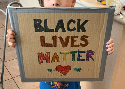 A young caucasian boy holds up a cardboard sign that reads Black Lives Matter with three cartoon hearts drawn below.  