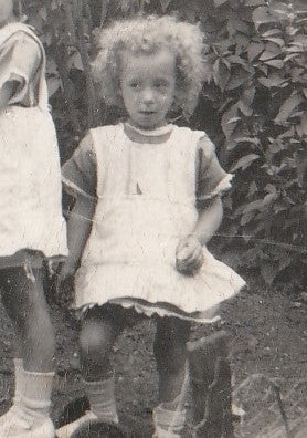 Image of a little curly haired girl in a dress and pinafore with bare legs, socks and shoes on.  Taken circa 1928.