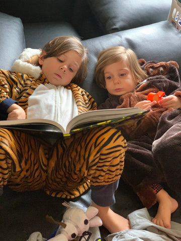 Two little boys lay on a sofa reading together.  one dressed as a tiger, one as a gorilla.