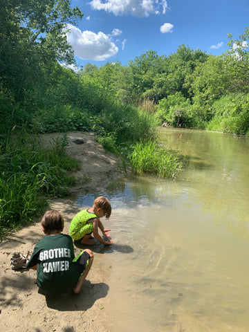 On the left, two small children play by a riverbank.  Behind them you can see the forest stretching in brilliant green with the blue sky above dotted with fluffy white clouds.  The river is murky to the bottom right of the image.  