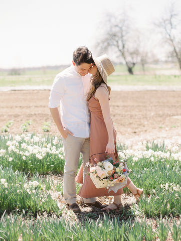 Couple kissing in a field of flowers with a spring bouquet in a basket and silk ribbon by The Lesser Bear Photo by Jenny Haas Florals Old Slate Farm Design and Planning Auburn and Ivory Creative