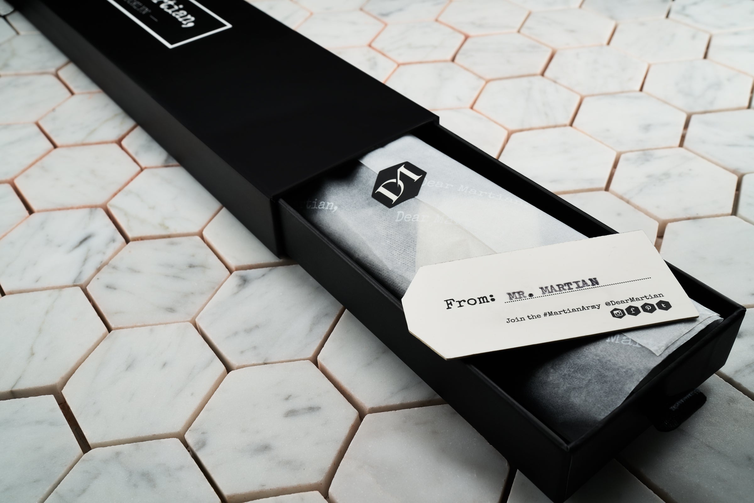 An image of Dear Martian, pull-out style box which contains a black and white checkered tie wrapped in DM gift wrapping. The wrapping is sealed with a DM hexagonal logo and in addition contains a personalized hang tag label with the Martian's name.