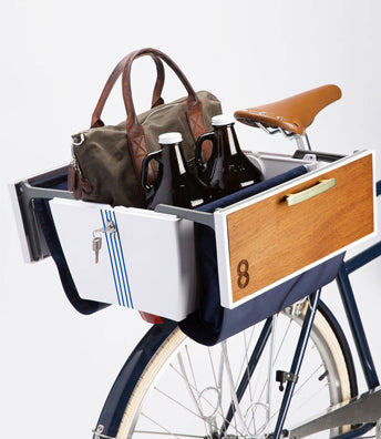 On a white background, the rear triangle and seat of a bicycle is visible. A rectangular locking bicycle trunk is installed on the rear rack. Trimmed with navy blue fabric and wood, the trunk is open to display the 2 growlers and weekend bag packed inside.