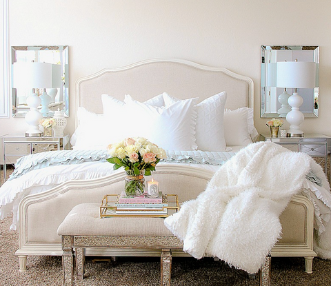 A beautiful king bed with ruffle bedding and a furry white throw, flowers on a bench