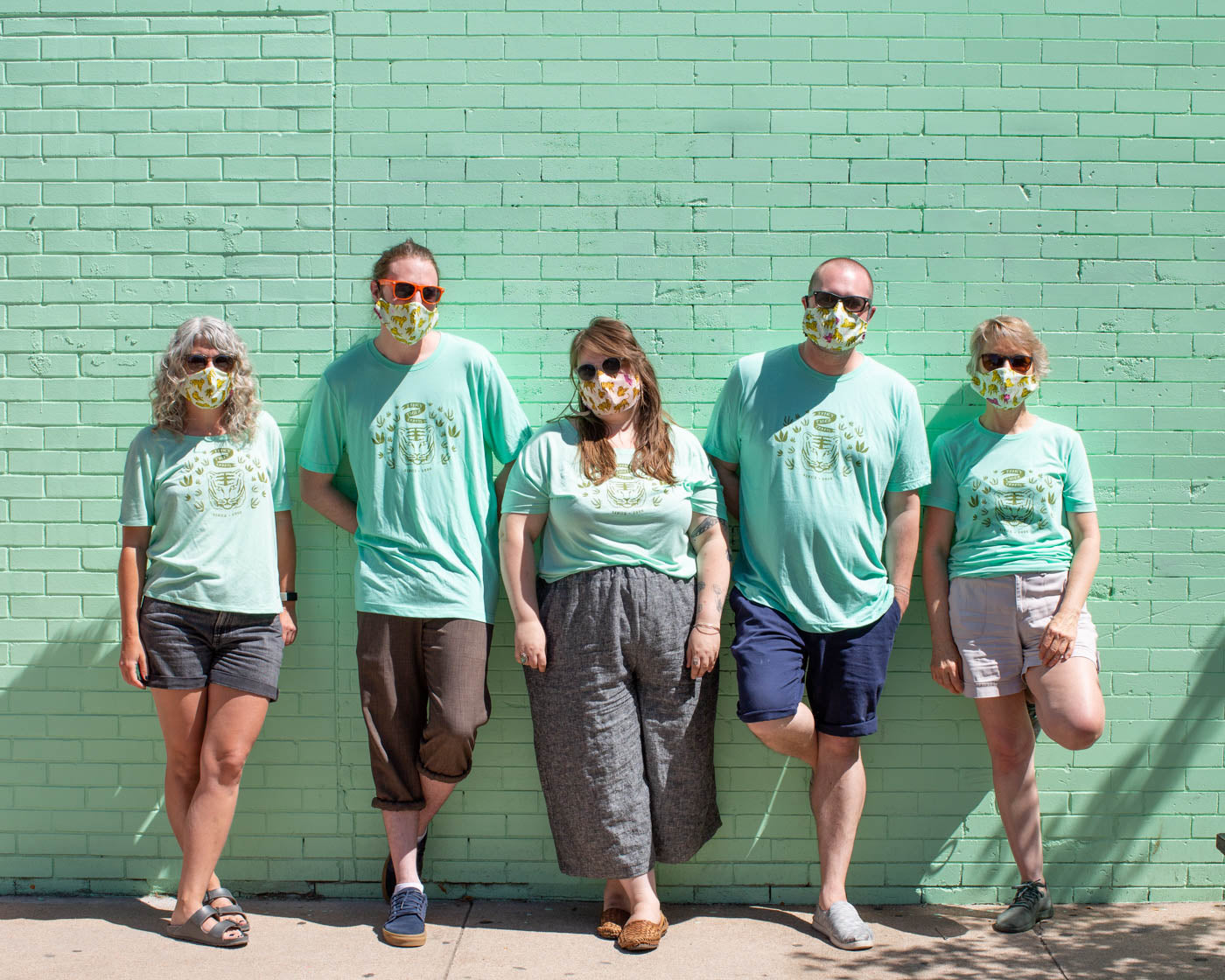 Five people, men and women, wearing matching mint green T-shirts against a mint green wall on a city sidewalk.