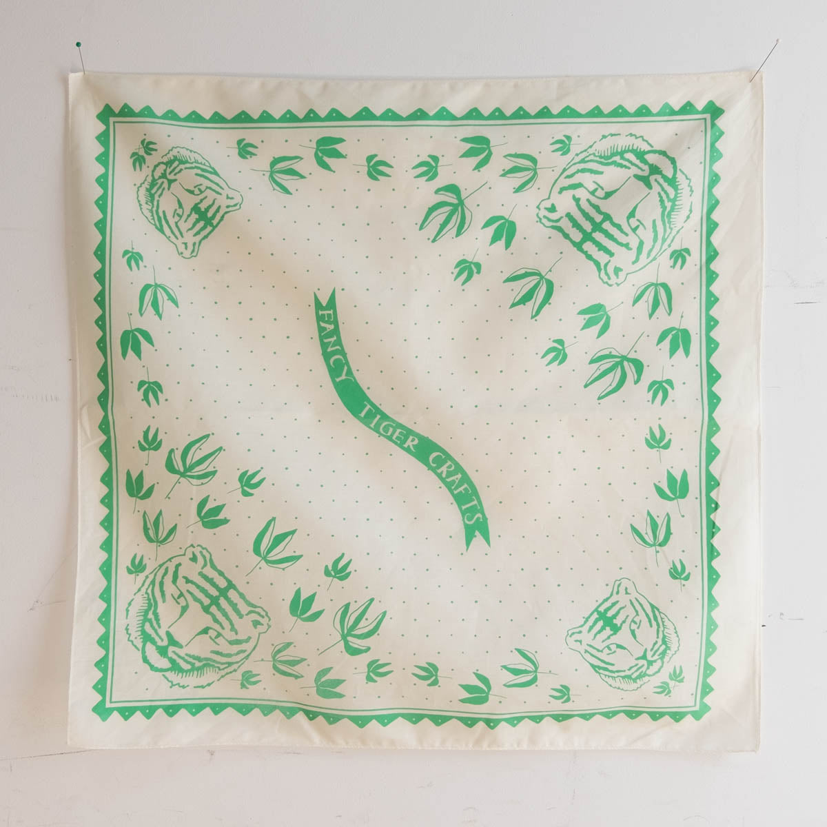 An off-white bandana, screenprinted in mint green, with a design featuring tiger faces and peony leaves. A banner that says "Fancy Tiger Crafts" is at the center of the design.  