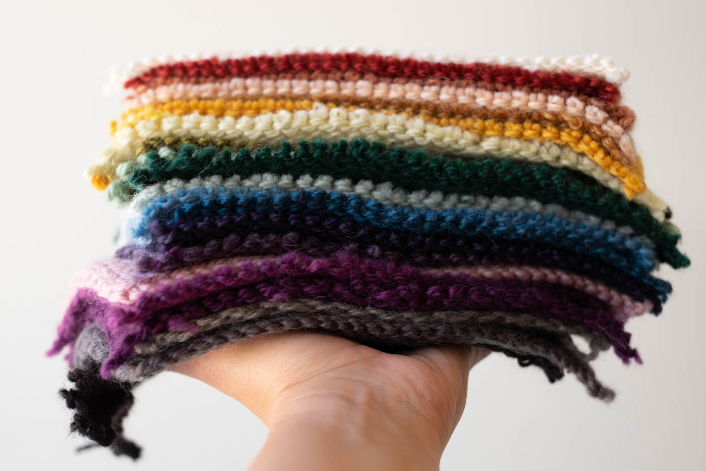 Yarn swatches stacked in a pile in a rainbow pattern being held by a hand straight out from the camera.
