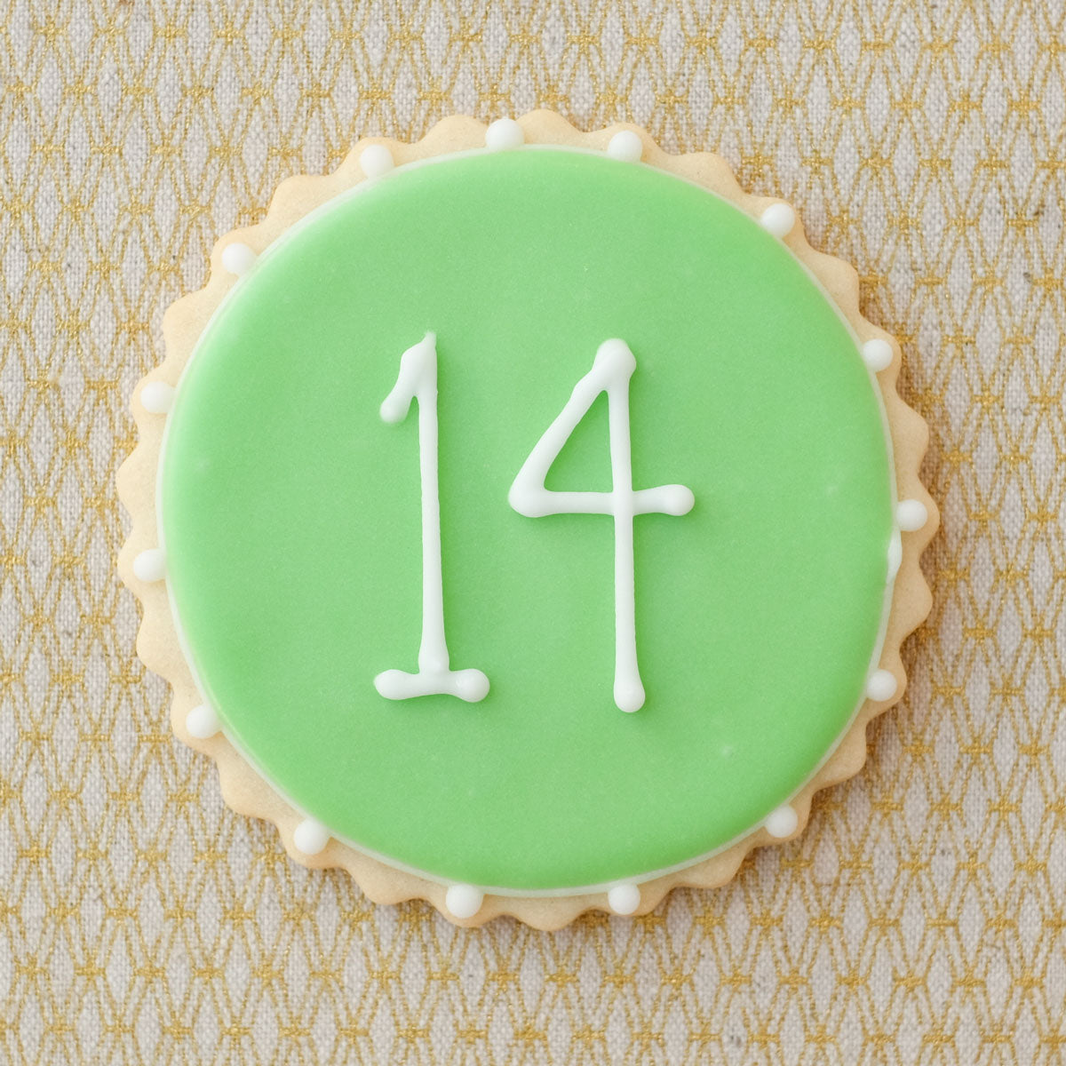 A round, scalloped edge cookie, frosted with mint royal icing and piped with the number 14.
