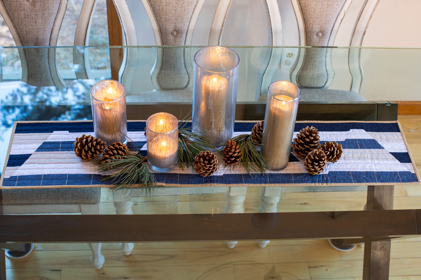 Photograph taken from above the glass kitchen table to showcase the table runner.  On top of the table runner sits lit candles, in tall candle holders, with pinecones and pine needle bunches intertwined.  