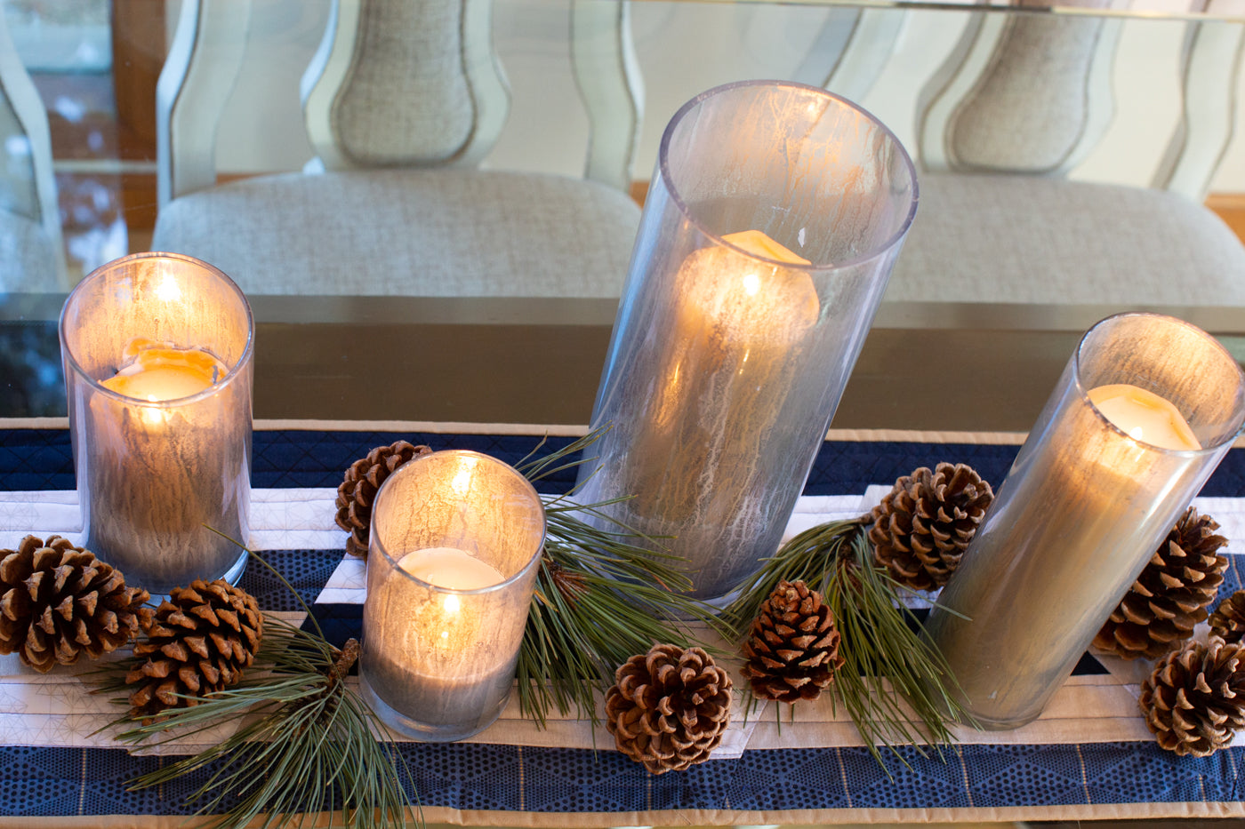 Photograph taken looking down at the kitchen table with a table runner.  The table runner is navy blue, white, tan and a deep blue.  There are candles lit in tall glass candle holders with pinecones and pine needle bunches at the base of the candles to create a holiday atmosphere.