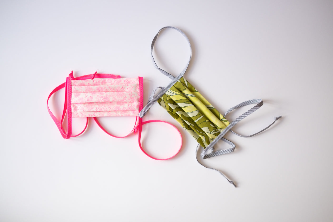 Face mask on a layflat on white background. Two face masks left face mask is bright pink and the right face mask is green.