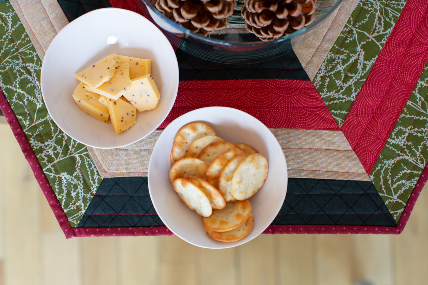 Photograph of looking at half of the hexagon centerpiece in Christmas green, red, a tan and black.  Two white small bowls are on the centerpiece one filled with sliced of mustard cheese and the other with white round crackers.