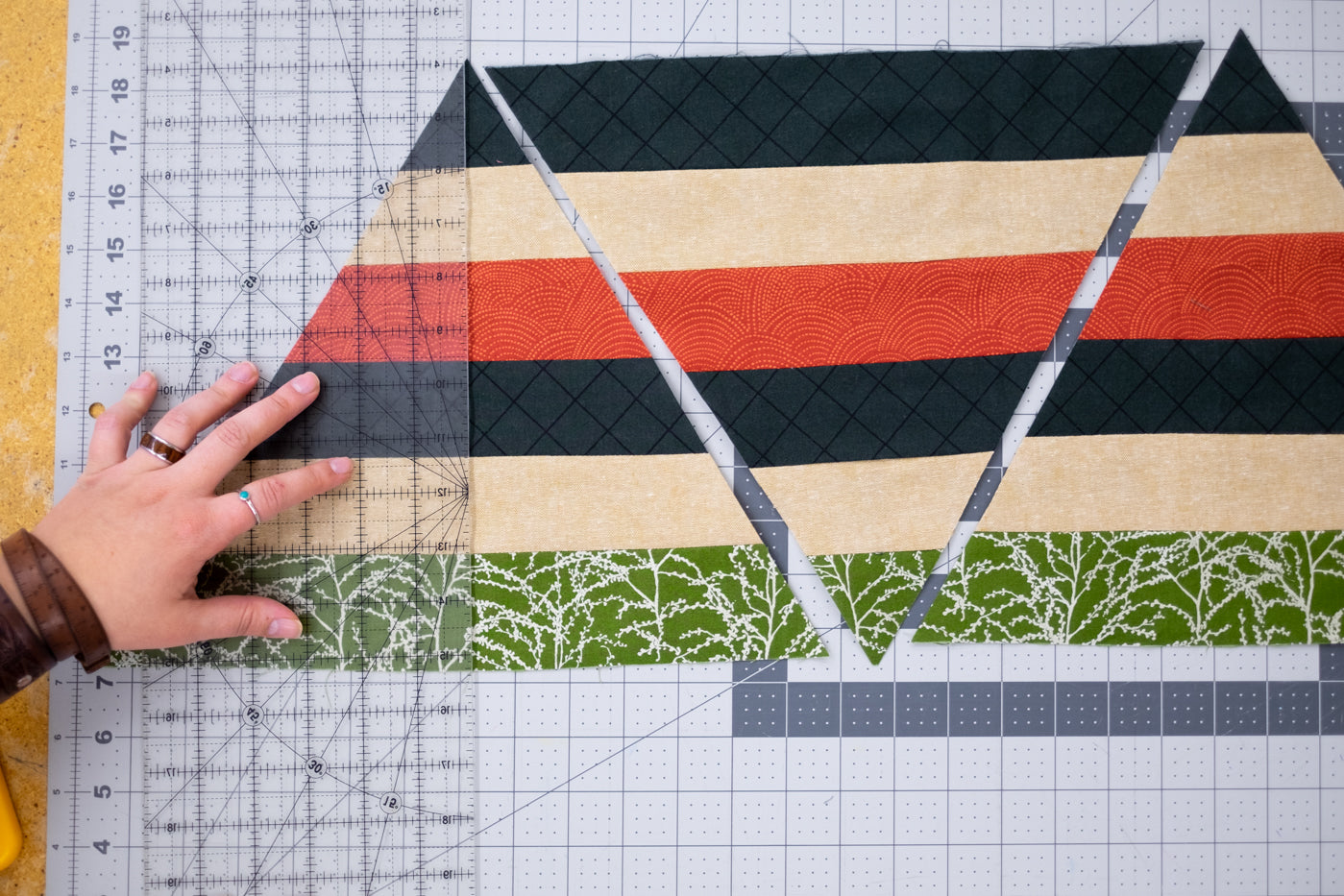 Material triangles laid out with a clear ruler over top showing them being lined up.  Triangles colors cut in lines of green with metallic sliver, soft yellow, black and red, sewn together to make a triangle. 